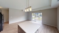 This thoughtfully designed single-floor unit offers a clean, contemporary aesthetic throughout. The heart of the home is an open-concept kitchen and living area, where light wood-tone flooring, neutral warm-gray walls, and abundant natural light create an inviting atmosphere. The kitchen is outfitted with sleek, textured dark cabinetry, white countertops, a central island with pendant lighting overhead, a double-basin sink, and black appliances including a range, microwave, and dishwasher. Track lighting provides additional illumination. The adjoining living and dining space flows seamlessly to a private balcony through sliding glass doors, offering outdoor access and views of the surrounding grounds. Two bathrooms serve the unit: a full bath featuring a tub-shower combination, wood-look vinyl flooring, chrome grab bars, and a white vanity with dark cabinetry; and a second bathroom with a large vanity mirror, bright vanity lighting, a white countertop, and dark wood-toned cabinets. The bedroom is carpeted for comfort, features a window with outdoor views, and includes a double-door closet. Throughout the unit, a cohesive palette of warm neutrals and modern finishes creates a polished, functional living environment.