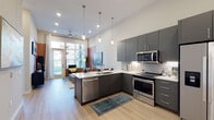 This single-floor unit showcases a cohesive modern design with light hardwood-style flooring and a neutral color palette accented by bold wall color-blocking throughout. The open-concept kitchen is a standout feature, equipped with stainless steel appliances including a French door refrigerator, electric range, built-in microwave, and dishwasher. Charcoal gray flat-front cabinetry reaches toward the ceiling, maximizing storage, while a white subway tile backsplash adds texture. A peninsula with a built-in sink and white quartz countertop extends into the living area, creating a natural transition between spaces. Pendant lighting overhead enhances the kitchen's contemporary feel. The living area benefits from floor-to-ceiling windows and a glass-paneled exterior door, drawing in abundant natural light and outdoor views. The bedroom continues the color-blocking design with a deep green accent wall, a large window with exterior views, and warm ambient lighting. The bathroom features a floating gray vanity with quartz countertop, a large mirror, tall storage tower, and light wood-style flooring, completing the unit's consistent modern aesthetic.