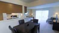 This single-floor residence showcases an open-concept design that seamlessly connects the dining and living areas, creating a cohesive and functional layout. The dining area is anchored by a dark-finish table paired with matching upholstered chairs, offering a defined gathering space within the open floor plan. Adjacent to the dining area, a pass-through window connects to the kitchen, which is partially visible and fitted with roller shades that complement the warm, neutral color palette throughout.

The living area extends toward a large sliding glass door dressed with vertical blinds, allowing generous natural light to fill the space and providing direct access to an outdoor area. The carpeted flooring adds warmth underfoot and contributes to the relaxed ambiance of the room. A sleeping area is visible in the background, furnished with a bed, bedside tables, and lamps, suggesting a studio or open-plan bedroom arrangement. Roller shades on the windows throughout the unit offer both privacy and light control. The overall aesthetic is clean, understated, and practical, with a consistent neutral tone unifying each zone of the space.