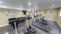 This fitness center offers a thoughtfully arranged workout space designed to accommodate a variety of exercise routines. The room features warm yellow-toned walls accented by an eye-catching motivational word-art mural, displaying fitness-related terms such as "Workout," "Energy," "Endurance," and "Strength," creating an energizing atmosphere for users.

The cardio section is well-stocked with multiple Star Trac treadmills and an elliptical trainer, providing ample options for cardiovascular exercise. A wall-mounted flat-screen television is positioned for easy viewing during workouts. A dedicated free weights area lines one wall, featuring a full rack of dumbbells alongside a weight bench, suitable for strength training routines.

The flooring combines hardwood-style surfaces with rubber mat sections beneath the equipment, offering both durability and safety. Recessed ceiling lighting provides bright, even illumination throughout the space. Natural light is introduced through a windowed door, contributing to the open and inviting feel of the room. Motivational framed artwork on the walls further reinforces the fitness-focused ambiance. Overall, this facility is clean, organized, and equipped to support a balanced fitness regimen.