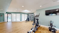 This single-floor fitness center is thoughtfully designed to provide a complete workout environment in a bright, welcoming setting. The space is finished with warm wood-look luxury vinyl plank flooring throughout, offering durability and a clean aesthetic underfoot. Walls are painted in a calming sage green palette, with bold motivational typography serving as a focal point on one accent wall, adding energy and character to the room.

Natural light pours in through a central skylight, complementing the recessed ceiling lighting and a ceiling fan that keeps the space comfortable during workouts. Large sliding glass doors and floor-to-ceiling roller shades line one wall, allowing for adjustable light control while providing views and direct access to an outdoor pool and lounge area beyond.

The fitness center is equipped with a range of cardio machines, including ellipticals, a treadmill, and a rowing machine, as well as a multi-cable strength training station paired with an adjustable bench. A full rack of dumbbells rounds out the free weight offerings, supporting a variety of strength training routines. A wall-mounted flat-screen television provides entertainment during workouts. A secondary entry door offers convenient access from an exterior walkway, making this fitness center easily accessible from multiple points within the property.