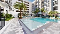 Located at 501 NW Seventh Ave in Fort Lauderdale, FL, this outdoor amenity space showcases a resort-style pool courtyard designed for relaxation and leisure. The centerpiece is a generously sized swimming pool with crystal-clear blue water, framed by sleek tiled pool decking in a sophisticated gray-and-white geometric pattern. Surrounding the pool, abundant tropical landscaping — including tall swaying palms, lush shrubs, and colorful plantings — creates a vibrant, resort-like atmosphere that reflects South Florida's natural character. A series of modern pergola structures with dark metal framing provide shaded seating areas outfitted with wicker lounge furniture, offering an inviting retreat from the sun. Rows of teal and aqua lounge chairs are positioned poolside beneath shade umbrellas for additional outdoor seating. The courtyard is enclosed by a sleek black iron safety fence and is flanked on multiple sides by a multi-story contemporary building with white facades, large windows, and private balconies that overlook the pool below. Glass-paneled entryways connect the outdoor space to the building's interior. The overall ambiance is modern, tropical, and refined — a well-appointed outdoor escape in the heart of Fort Lauderdale.