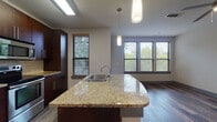 This single-floor unit is designed with a contemporary, open-concept layout that integrates the kitchen and living areas into one cohesive space. The kitchen serves as a central feature, showcasing a large granite-topped island with a stainless steel sink and elegant pendant lighting overhead. Rich, dark-toned cabinetry lines the walls, complemented by frosted glass upper cabinet inserts, a subway tile backsplash, and a full suite of stainless steel appliances including a side-by-side refrigerator, electric range, and built-in microwave. Dark hardwood-style flooring extends throughout the kitchen and living area, adding warmth and continuity. Multiple windows with horizontal blinds invite natural light and offer views of surrounding greenery, enhancing the overall brightness of the space. A door with a full-length sidelight panel provides additional natural light and outdoor access. The bathroom features large-format tile flooring, a tiled tub-shower combination with recessed lighting, and a dark wood vanity. The bedroom is carpeted and finished with neutral tones, a ceiling fan, and ample wall space, offering a comfortable, quiet retreat within the unit.