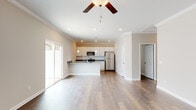 This well-appointed single-story home showcases a thoughtfully designed open-concept layout that seamlessly connects the kitchen and living areas. The kitchen serves as a central hub, featuring granite countertops, white shaker-style cabinetry, a stainless steel undermount sink set within a peninsula island, and pendant lighting overhead. Stainless steel appliances — including a range, microwave, and refrigerator — are complemented by a classic white subway tile backsplash. The living area extends naturally from the kitchen, bathed in natural light from a sliding glass door that opens to an outdoor view. A ceiling fan anchors the living space, which is finished with warm hardwood-style flooring that flows throughout the home. The bedrooms are generously proportioned with ceiling fans, natural light from windows, and rich dark hardwood-style floors. The primary suite includes a well-sized bathroom with granite-topped dual vanity, white cabinetry, and a dedicated walk-in closet with built-in wire shelving. A utility area houses a commercial-grade water heater, adding to the home's practical infrastructure. Crown molding and recessed lighting throughout lend a refined, cohesive aesthetic to this move-in-ready residence.