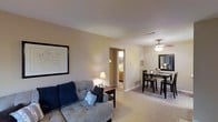 This single-floor residence offers a thoughtfully arranged layout that maximizes both comfort and functionality. The living area serves as the heart of the space, featuring plush, light-toned carpeting underfoot and neutral-painted walls that create a warm, inviting atmosphere. A cozy sofa with decorative pillows anchors the room, accompanied by a side table and accented by framed wall art that adds a personal touch. Recessed ventilation and a ceiling fan contribute to year-round comfort throughout the space. The living area flows seamlessly into a dedicated dining space, where a dark-toned dining table and matching chairs sit beneath a ceiling fan with integrated lighting, making it an ideal setting for everyday meals. A decorative room divider with a lattice-style design provides a subtle yet stylish separation between the two areas. A large mirror in the dining space enhances the sense of openness. Adjacent to the main living area, a hallway leads to what appears to be a well-appointed bathroom, visible in the background, featuring warm cabinetry and a bright vanity area. The overall ambiance of the space is calm and welcoming, with consistent neutral tones and warm lighting tying each area together cohesively.