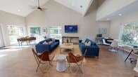 Located at 66 Daughtrey Ave in Waco, Texas, this single-floor space delivers a bright, modern aesthetic defined by dramatic vaulted ceilings and an open-concept layout. The expansive main living area serves as the heart of the space, anchored by bold blue sofas, a woven jute rug, and warm wood-look flooring that flows seamlessly throughout. A wall-mounted flat-screen TV, ceiling fan, and an abundance of recessed lighting make the area both functional and inviting. Large windows and glass doors flood the interior with natural light while offering views of an outdoor pool and deck area. A dedicated workspace features a rustic wood desk positioned beneath oversized windows, providing an inspiring, light-filled environment. The kitchenette is thoughtfully appointed with white shaker cabinetry, dark countertops, a stainless steel refrigerator, a subway tile backsplash, and a round gold-framed mirror as a decorative focal point. A built-in banquette nook with a round white table adds a charming and practical dining area. A cozy lounge nook near the entry offers two accent chairs bathed in sunlight from a large glass door that opens directly to the outdoor deck. The overall ambiance is clean, contemporary, and welcoming, blending functional design with warm, stylish finishes.