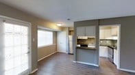 This single-floor space offers an open and functional layout that seamlessly connects the living area to the kitchen. The living area is bathed in natural light through a large window fitted with white horizontal blinds, complementing the warm, medium-toned wood-look flooring that runs throughout the space. Neutral beige walls provide a welcoming backdrop, while white baseboards offer a clean contrast.

The kitchen is distinctly defined by a gray accent wall and peninsula-style counter with dark countertops, creating a natural division between cooking and living spaces. Upper cabinetry is finished in gray and white, adding a modern, two-tone aesthetic. A mosaic tile backsplash in earthy tones adds texture and visual interest behind the sink and stovetop area. Stainless steel appliances, including a refrigerator, contribute to the kitchen's contemporary feel. A second set of white cabinetry lines an adjacent wall, providing ample storage. The textured ceiling adds a subtle design element overhead. A doorway visible from the main area hints at additional rooms beyond, suggesting a practical and well-organized floor plan overall.