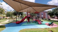 This single-story community clubhouse offers a thoughtfully designed gathering space that blends sophisticated interiors with practical recreational amenities. The entrance features rich dark-wood-framed glass doors and travertine tile flooring that carries throughout the interior, establishing a warm, upscale tone from the moment of arrival.

The main lounge area is anchored by a stone-clad fireplace surround and tray ceiling with recessed lighting, creating an inviting focal point. Elegantly patterned wingback chairs and dark wood accent tables furnish the space, while interior French doors with glass panels connect adjacent rooms, maintaining an open and airy feel. A kitchenette or bar area is visible toward the rear of the lounge.

A dedicated game room accommodates a full-size billiards table and a shuffleboard table, illuminated by pendant lighting. High windows along one wall flood the space with natural light and offer views of the surrounding landscaped grounds. Wall sconces and framed artwork contribute to the polished atmosphere throughout.

Outdoors, a generously sized covered playground sits beneath a large tensile shade canopy, featuring a multi-component play structure with slides, climbing elements, and rubber safety surfacing in bold colors, nestled within a well-maintained green lawn adjacent to brick residential buildings.