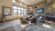This single-floor community lounge is designed to impress from the moment you enter. Soaring ceilings with clerestory windows flood the space with natural light, creating an open and airy atmosphere throughout. The focal point of the main lounge area is a striking floor-to-ceiling stacked stone fireplace, complemented by a dark wood mantel and a large mounted flat-screen television on an adjacent textured accent wall. Comfortable gray upholstered sofas and chairs are arranged around a round wood coffee table atop a patterned area rug, inviting relaxed conversation and gathering.

A reception counter with a dark wood base and granite surface marks a transition into a secondary area, which features a casual dining nook with a round table and upholstered chairs, bathed in light from large floor-to-ceiling windows overlooking the surrounding landscape. Patterned carpet tiles in neutral tones of gray and taupe flow throughout, adding visual texture underfoot. Pendant lighting and a ceiling fan provide both ambiance and function across the space.

The overall design blends contemporary comfort with traditional warmth, making this a versatile and welcoming communal space suited for both social gatherings and everyday use.