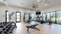 Located at Yardly Artisan Lakes in Palmetto, Florida, this dedicated fitness center offers a thoughtfully designed single-room workout space that accommodates a comprehensive range of exercise options. The room features light hardwood-style flooring, crisp white walls, and recessed lighting complemented by large ceiling fans that promote air circulation throughout the space.

Natural light pours in through multiple large windows fitted with sleek black frames, offering views of the surrounding Florida landscape, including palm trees that reinforce the community's tropical setting. A wall-mounted flat-screen television provides entertainment during workouts.

The fitness center is outfitted with a robust selection of equipment to support both cardio and strength training. Cardio options include treadmills, an elliptical, and a stationary bike, all featuring integrated digital displays. Strength training equipment includes a power rack with weight plates, a cable machine, and a multi-function functional trainer. Adjustable weight benches and a full dumbbell rack line one wall, while a dedicated storage rack holds medicine balls, kettlebells, and stability balls for functional fitness workouts. A rowing machine rounds out the cardio offerings. The space is clean, modern, and efficiently organized, making it a well-equipped amenity for residents seeking a convenient on-site fitness option.
