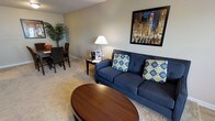 This single-floor residence offers a welcoming and well-appointed layout designed for comfortable everyday living. The open-concept floor plan seamlessly connects the living and dining areas, creating a cohesive and functional space that feels both inviting and practical.

The living area is centered around a plush navy-blue sofa accented with vibrant geometric throw pillows, complemented by a rich, oval wood coffee table that anchors the room. A classic table lamp on a side table provides warm ambient lighting, while a bold cityscape artwork displayed prominently on the wall adds a dynamic focal point and a touch of urban character to the space. Soft, neutral-toned wall-to-wall carpeting runs throughout, contributing to the room's comfortable and relaxed atmosphere.

Adjacent to the living area, the dining space features a dark wood dining table and chairs, ideal for meals and gatherings. A lush indoor tropical plant adds a refreshing natural element, enhancing the overall warmth of the space.

The neutral wall tones throughout provide a versatile backdrop that ties the living and dining zones together harmoniously. The layout is efficient and thoughtfully arranged, making excellent use of the available space while maintaining an open, airy feel. Overall, this residence offers a clean, move-in-ready environment with a cozy and functional ambiance.