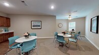 Located at 201 Kroger Drive in Richmond Hill, Georgia, this single-floor community space is thoughtfully designed to foster social interaction and comfort. The main gathering area features soft, neutral-toned walls paired with plush carpeted flooring, creating a warm and inviting atmosphere. Multiple round white tables, each surrounded by contemporary teal chairs, are arranged throughout the room to encourage conversation and group dining. A ceiling fan provides additional comfort, while natural light filters in through a window with blinds, adding brightness to the space. Coastal-themed artwork, including framed depictions of herons and seascapes, lends the room a cohesive, regionally inspired aesthetic. The kitchen area transitions seamlessly from the gathering space and is outfitted with rich, medium-toned wood cabinetry extending across the full wall, complemented by clean white countertops and a double stainless steel sink. Durable hardwood-style flooring grounds the kitchen, offering a practical and attractive contrast to the carpeted lounge area. Glass-paneled interior doors connect the spaces, enhancing visibility and flow. The overall design balances functionality with a relaxed, welcoming ambiance suited for community gatherings and shared activities.
