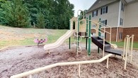 Jefferson is an outdoor community space designed to serve as a recreational hub for residents. The centerpiece of this area is a well-equipped playground featuring a multi-level play structure with slides, climbing elements, and accessible ramps, all finished in neutral tones complemented by green support poles. The ground is covered with wood chip mulch, providing a safe and cushioned surface for active play. A pink spring rider adds a playful touch to the area, offering additional entertainment options for younger visitors. The playground is bordered by a gently sloping, tree-lined hillside filled with mature deciduous trees, creating a natural green buffer that enhances the sense of privacy and connection to the outdoors. Adjacent to the play area stands a brick residential building with classic architectural details, including black shutters and multiple windows, reinforcing the community-oriented character of the space. The overall ambiance is open, family-friendly, and nestled within a natural setting that balances structured amenities with the charm of the surrounding landscape.