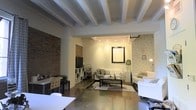 This single-floor space masterfully combines industrial character with contemporary design. The open-plan layout is anchored by striking exposed brick walls that add warmth and texture, contrasting beautifully with the crisp white painted beam ceilings above. The beamed ceiling detail lends architectural depth and a loft-like quality to the interior.

The space is divided into functional zones that flow seamlessly into one another. A comfortable lounge area features neutral-toned sofas arranged around a coffee table, grounded by a patterned area rug. Subtle patterned wallpaper adds visual interest to one section of the walls, softening the industrial aesthetic. Track lighting installed along the ceiling provides adjustable, targeted illumination throughout, complementing the warm ambient glow of floor and table lamps.

A dedicated work or dining area is positioned toward the front, furnished with a white table and chairs, ideal for collaborative or individual use. Storage is thoughtfully integrated with open shelving units and cabinetry. The polished concrete floors tie the entire space together, offering a sleek, durable surface that reinforces the industrial-modern character. Overall, the space exudes a versatile, well-curated ambiance suitable for both productive work and relaxed gatherings.