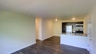 This single-floor residence offers a practical and well-organized open-concept layout that seamlessly connects the living area and kitchen. The space is anchored by rich, dark hardwood-style flooring that contrasts elegantly with the light, neutral-toned walls, creating a clean and inviting atmosphere throughout.

The kitchen is a standout feature, equipped with dark cabinetry that provides ample storage and a striking visual contrast against the lighter surroundings. A peninsula with a granite-style countertop defines the kitchen boundary, offering additional prep space and a natural transition into the open living area. A white refrigerator and integrated sink round out the functional kitchen setup.

The living area is generously proportioned, providing flexible space for a variety of furniture arrangements. The textured ceiling adds a subtle architectural detail, while flush-mount ceiling fixtures provide consistent overhead lighting throughout. A paneled interior door leads to additional rooms, suggesting further functional spaces beyond the main open area.

Overall, the residence presents a cohesive, move-in-ready environment with a neutral palette and durable finishes that balance style and practicality, making it a versatile and comfortable living space.