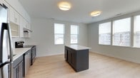 This single-floor residence offers three bedrooms and two bathrooms within a thoughtfully designed open-concept layout. The kitchen and living area form the heart of the home, highlighted by a sleek dark-cabinetry island with a crisp white countertop that serves as both a functional workspace and a natural gathering point. Light hardwood flooring extends throughout, lending warmth and continuity to the space. Multiple windows dressed with white horizontal blinds flood the interior with natural light while framing views of the surrounding neighborhood. The kitchen is equipped with dark appliances that complement the island's cabinetry, creating a cohesive, contemporary aesthetic. Neutral gray-toned walls provide a clean, versatile backdrop that suits a variety of furnishing styles. Flush-mount ceiling fixtures provide consistent overhead illumination, reinforcing the bright and airy atmosphere. The single-floor design ensures ease of accessibility and a seamless flow between living spaces. Overall, the home presents a modern, functional aesthetic with clean lines and quality finishes throughout.