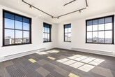 This single-floor unit combines industrial-modern design with functional living spaces. The main living area is a standout feature, characterized by expansive black-framed grid windows that flood the room with natural light and frame elevated city views. Track lighting runs along black pipe conduits across the white ceiling, adding an industrial aesthetic, while patterned carpet tiles in grey, tan, and yellow tones cover the floor throughout the living and sleeping areas.

The kitchen is well-appointed with light wood cabinetry, dark granite countertops, and stainless steel appliances including a refrigerator, electric range, and microwave. A peninsula with an integrated sink provides additional workspace and casual seating potential. Pendant lighting and warm flooring give this area a welcoming feel.

The bedroom offers a comfortable layout with the same patterned carpet, a sliding closet with ample storage, and light wood doors consistent with the unit's design language. A window provides additional natural light and city views.

The bathroom features distinctive mosaic tile flooring in a geometric pattern, a full tub-shower combination, and the added convenience of a stacked washer and dryer unit tucked neatly within the space. Clean white walls and modern fixtures complete the room.