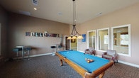 This single-floor recreational space is designed as a dedicated game and lounge room, centered around a full-size billiards table with a vibrant blue felt surface and elegant carved wood legs. A pendant light fixture with warm-toned shades hangs directly above the table, providing focused illumination ideal for gameplay. The room features neutral warm-beige walls and patterned carpeting throughout, contributing to a casual yet polished atmosphere.

Along one wall, a "BILLIARDS" letter display and mounted cue rack add a playful, thematic touch. Multiple windows along the exterior wall allow natural light to filter in, with views of the surrounding outdoor area. A wall-mounted flat-screen television is positioned for easy viewing from the lounge area, which includes bar-height tables with stools and small accent tables with seating.

A cozy nook within the space features two upholstered chairs flanking a granite-topped accent table, decorated with artistic wall mirrors and a vintage-style game clock, adding character to the room. Additional lounge chairs are positioned near the billiards area, making this space versatile for both active play and relaxed socializing. The overall layout is open and well-organized, suitable for community or shared recreational use.