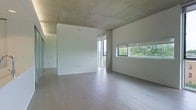This single-floor space showcases a compelling blend of industrial and modern design elements. The exposed concrete ceiling serves as a defining architectural feature, adding raw texture and visual interest that contrasts with the clean, white walls below. Light-toned hardwood flooring runs throughout, contributing warmth and a sense of continuity across the open layout.

Natural light is a prominent characteristic of this space, introduced through two distinct window configurations. A tall, floor-to-ceiling window on one wall allows light to pour in vertically, while a wide horizontal window on the adjacent wall frames lush, green tree canopy views, connecting the interior to the surrounding landscape. Together, these openings ensure the space remains bright and airy throughout the day.

The open floor plan offers considerable flexibility, with no interior partitions interrupting the flow of the main area. The neutral color palette — spanning white walls, pale flooring, and grey concrete — creates a calm, versatile backdrop well-suited to a variety of uses. The overall ambiance is minimal, modern, and understated, emphasizing clean lines and quality materials over ornamentation.