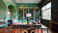 This single-story space makes a strong design statement through its bold use of jewel-toned color, blending deep teal, rich emerald green, and warm wood tones throughout. The entry foyer welcomes visitors with dark teal walls, tall black-framed glass doors, patterned area rugs over wide-plank hardwood floors, and potted greenery that adds a natural touch.

Moving inward, the open main hall features expansive hardwood flooring, teal-painted walls adorned with curated artwork, and a central bar-height counter topped with light wood stools set against a dramatic arched, wallpapered accent niche with a gold circular mirror. Pendant lighting in brass and geometric styles is distributed throughout, contributing to a warm, sophisticated ambiance.

A library lounge area is defined by floor-to-ceiling built-in bookshelves painted emerald green, filled with books and decorative objects, flanked by upholstered armchairs and anchored by a central desk. An arched alcove with built-in bench seating adds architectural interest. Large windows throughout the space admit generous natural light and offer views to the surrounding streetscape.

Private meeting areas enclosed by sleek black-framed glass partitions provide functional separation while maintaining visual openness. A long communal dining table with black chairs occupies a dedicated gathering zone. The overall atmosphere is curated, eclectic, and inviting.