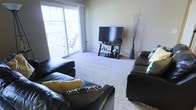 This single-floor space is centered around a welcoming living room that combines comfort and practicality. The room is finished with plush wall-to-wall carpeting in a neutral tone, contributing to a warm and relaxed ambiance throughout. A large sliding glass door floods the space with natural light and provides direct access to an outdoor balcony, where outdoor seating can be seen, offering a pleasant extension of the living area. The neutral-toned walls provide a clean, versatile backdrop that complements a range of furnishings and décor styles. The living room is arranged around dark leather sofas accented with decorative yellow and patterned cushions, creating a cozy seating area. A flat-screen television is positioned on a glass and metal media stand along one wall, making this a functional entertainment space. A tall decorative floor vase with branching accents adds an artistic touch to the corner of the room. The overall layout is open and uncluttered, allowing for easy movement throughout the space. The combination of natural light, soft carpeting, and direct outdoor access gives this space a comfortable, livable character suited to everyday relaxation and entertaining.