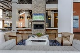 This expansive single-floor space showcases a striking blend of industrial and contemporary design elements. Soaring ceilings with exposed ductwork and structural beams lend an open, loft-like atmosphere, while wood-look tile flooring flows seamlessly throughout, unifying the various functional zones.

The kitchen is a standout feature, equipped with stainless steel appliances including a professional-grade range and French-door refrigerator. Dark cabinetry contrasts beautifully with granite countertops and an ornate patterned tile island surround, while a dark subway tile backsplash and bold pendant lighting complete the sophisticated aesthetic.

Multiple lounge areas are thoughtfully distributed across the floor, each with its own distinct character. One features a stone-clad fireplace column with a wall-mounted television, flanked by neutral sofas and accent seating. Another seating vignette incorporates a rich navy sofa and leather chairs atop a geometric area rug. A dining area with upholstered chairs and patterned carpet sits adjacent to warm terracotta-toned walls adorned with curated artwork.

Decorative touches, including large sculptural leaf wall art and warm pendant lighting, add warmth and personality to the space, creating an inviting, well-appointed environment suited for gathering and relaxation.
