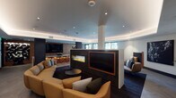 This upscale amenity lounge is designed with a refined contemporary aesthetic, offering a thoughtfully arranged layout that balances social gathering and intimate relaxation. The centerpiece of the space is a striking double-sided linear fireplace set within a dark, textured partition wall, which also houses a large flat-screen TV framed in warm wood trim — elegantly dividing the room into distinct seating areas. Curved, tan leather sofas anchor the main lounge zone atop a deep navy area rug, complemented by sleek, minimalist coffee tables. Cove lighting runs along the perimeter of the tray ceiling, casting a warm, ambient glow that enhances the polished atmosphere. A built-in window nook provides an additional casual seating option alongside large windows that bring in natural light and views of the surrounding exterior. The kitchenette bar area features a pristine white stone countertop with an under-mount sink, warm wood wall paneling, integrated display shelving, and a wall-mounted TV — ideal for social gatherings. Throughout the space, curated artwork, lush potted greenery, and carefully selected furnishings in a rich palette of mustard, tan, and dark tones contribute to a cohesive, hotel-caliber environment.