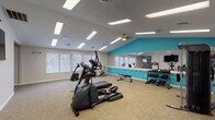 This fitness center is organized across a single floor into several distinct workout zones, offering a comprehensive exercise environment. The main cardio area is generously sized, featuring treadmills, elliptical trainers, and a stationary bike arranged on durable carpet tile flooring. A wall-mounted flat-screen TV provides entertainment during workouts, while multiple windows with blinds allow natural light to fill the space. Recessed panel lighting and a vaulted ceiling section with a ceiling fan add to the room's open, airy feel. A full-length mirror wall with a teal accent wall enhances the sense of depth and energizes the aesthetic. An adjoining strength training section houses an Inspire cable machine system, offering versatile resistance training options. A dedicated functional training room provides space for floor-based exercises, equipped with stability balls, resistance tools, wall-mounted exercise guide posters, and a mirrored wall. This room also features a ceiling fan and its own window for ventilation and light. A dumbbell rack with an organized free weight selection rounds out the strength training offerings. A corridor with glass partition walls connects the various areas, providing a clean and accessible flow throughout the facility. The overall design is modern and functional, suited for a full-body fitness routine.
