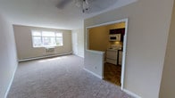 Located at 17 W Chester Pike in Ridley Park, Pennsylvania, this single-floor unit offers a straightforward and functional layout designed for comfortable everyday living. The living area is finished with wall-to-wall carpeting that adds warmth underfoot, and is illuminated by a ceiling fan light fixture overhead. A window-mounted air conditioning unit and baseboard heating ensure year-round climate control. Natural light filters in through a set of windows along the exterior wall, creating a bright and welcoming atmosphere. An open passthrough connects the living area to the kitchen, which features dark wood cabinetry, a white electric range, built-in microwave, and tile flooring in a classic pattern. The kitchen is compact yet well-equipped, offering an efficient workspace. A white interior door in the living area provides access to additional areas of the unit. The neutral color palette throughout — from the beige walls to the light carpet — gives the space a clean, move-in-ready feel that is easy to personalize. Overall, this unit at 17 W Chester Pike presents a practical and comfortable living environment in the Ridley Park community.
