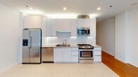 This single-floor residence presents a clean, contemporary design characterized by an open layout and modern finishes throughout. The main living area is bright and airy, featuring warm hardwood flooring and crisp white walls. Floor-to-ceiling sliding glass doors lead to a private balcony, framing urban cityscape views and drawing natural light deep into the interior. A wall-mounted split air conditioning unit ensures year-round comfort.

The kitchen is seamlessly integrated into the living space and is outfitted with flat-front white cabinetry, stone countertops, and a classic subway tile backsplash. Stainless steel appliances — including a gas range, built-in microwave, dishwasher, and refrigerator — complete the space, which transitions from tile flooring into the hardwood of the living area.

The bathroom is finished to a high standard, featuring a marble-patterned stone vanity countertop with an undermount sink, dark wood cabinetry, and a frameless sliding glass shower enclosure with textured white tile surrounds. Recessed lighting and a large mirror contribute to the clean, polished aesthetic. Overall, the unit offers a cohesive modern ambiance throughout.