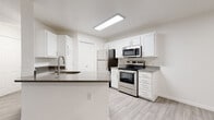 This single-story space features a clean, modern design with a consistent neutral palette and light wood-look flooring carried throughout every room. The kitchen serves as a functional centerpiece, showcasing white shaker-style cabinetry extending to the ceiling for generous storage, contrasted by dark countertops that add visual depth. A peninsula with an undermount sink provides additional workspace and a natural division between the kitchen and the adjoining living area. Stainless steel appliances, including a refrigerator, electric range, and over-the-range microwave, complete the well-equipped cooking space. Overhead flush lighting keeps the kitchen bright and practical. The living and bedroom areas are finished with the same light wood-look flooring and crisp white walls, lending a clean, open feel. Windows fitted with horizontal blinds allow natural light to filter in while maintaining privacy. The bathrooms mirror the home's neutral aesthetic, featuring white tub-shower combinations with curved curtain rods, white vanity cabinetry with dark countertops, and the same wood-look flooring. Brushed nickel and gold-toned hardware details are present throughout, providing subtle warmth against the predominantly white and grey interior.