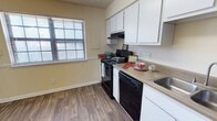 This kitchen space showcases a functional and contemporary design. The room is outfitted with a full suite of black appliances, including a stove, microwave, and dishwasher, creating a cohesive look against the white cabinetry. The cabinets extend both above and below the countertop, offering abundant storage options. A light-colored countertop provides ample workspace for meal preparation.

The floor features wood-look laminate or vinyl planks in a warm, medium tone, adding warmth and durability to the space. A window with horizontal blinds allows natural light to filter in, brightening the room and offering a view to the outside. The walls are painted in a soft, neutral color, complementing the overall color scheme.

Colorful dishware and decorative items on the counter add pops of color to the predominantly neutral palette. The layout appears to be galley-style, maximizing efficiency in a compact area. Overall, this kitchen combines practicality with a clean, modern aesthetic, suitable for everyday use in an apartment or small home setting.
