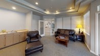 Located at 901 Sunburst Ct in Moses Lake, Washington, this single-floor community clubhouse offers a thoughtfully designed gathering space with a range of amenities to support both relaxation and an active lifestyle. Throughout the facility, tray ceilings with recessed lighting lend an elegant, polished feel, while neutral tones and carpeted floors create a warm and welcoming atmosphere. The fitness center is a standout feature, divided into two well-equipped zones. One area houses a treadmill, recumbent bike, and upright cycle beneath a tray ceiling with motivational wall decals and ample natural light. An adjacent room contains an elliptical trainer and a stepper machine, with direct access to the outdoors through a glass door. The lounge area provides a comfortable retreat, furnished with leather seating, a coffee table, and a media console with built-in shelving — ideal for residents to unwind or socialize. A dedicated dining room offers seating for a large group around a rich wood table, surrounded by windows fitted with blinds that fill the space with diffused natural light. The layout flows cohesively between spaces, making this clubhouse a functional and inviting hub for the community.