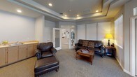 Located at 901 Sunburst Ct in Moses Lake, Washington, this single-floor community clubhouse offers a thoughtfully designed gathering space with a range of amenities to support both relaxation and an active lifestyle. Throughout the facility, tray ceilings with recessed lighting lend an elegant, polished feel, while neutral tones and carpeted floors create a warm and welcoming atmosphere. The fitness center is a standout feature, divided into two well-equipped zones. One area houses a treadmill, recumbent bike, and upright cycle beneath a tray ceiling with motivational wall decals and ample natural light. An adjacent room contains an elliptical trainer and a stepper machine, with direct access to the outdoors through a glass door. The lounge area provides a comfortable retreat, furnished with leather seating, a coffee table, and a media console with built-in shelving — ideal for residents to unwind or socialize. A dedicated dining room offers seating for a large group around a rich wood table, surrounded by windows fitted with blinds that fill the space with diffused natural light. The layout flows cohesively between spaces, making this clubhouse a functional and inviting hub for the community.