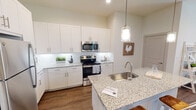 Located in Waxahachie, TX, this single-story home blends contemporary design with warm farmhouse-inspired touches throughout. The kitchen serves as a central highlight, showcasing white shaker cabinetry, granite countertops, a subway tile backsplash, stainless steel appliances, and a large island with an undermount sink and pendant lighting overhead. Wood-look flooring runs throughout the main living areas, adding warmth and continuity to the space. Adjacent to the kitchen, a built-in desk nook with granite countertop and open shelving offers a practical workspace, while a nearby dining area provides an intimate setting for meals. The living room is bright and airy, benefiting from multiple windows that invite generous natural light, and features elevated ceilings that lend a sense of spaciousness. The bedroom is generously sized, accommodating a four-poster canopy bed frame and a large window with ample natural light and ceiling fan. The bathroom features a tub-shower combination elegantly clad in large-format marble-look tile, creating a clean and refined aesthetic. Throughout the home, a neutral color palette and consistent finishes create a cohesive, modern atmosphere with a welcoming, lived-in charm.