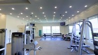This fitness center is a comprehensive, single-level workout facility designed to accommodate a wide range of exercise needs. The space is illuminated by an abundance of recessed ceiling lights, ensuring a bright and energizing environment throughout. Large windows along one wall invite natural light and provide open views of the surrounding area, contributing to an airy and motivating atmosphere.

The facility is outfitted with a robust selection of professional-grade equipment. Precor strength machines anchor one side of the floor, offering a variety of resistance training options. A full dumbbell rack lines the far wall, featuring an extensive range of free weights neatly organized for easy access, accompanied by exercise benches positioned on protective floor mats. A cable and multi-station weight machine further expands the strength training options available to users. Additional functional training equipment, including medicine balls, rounds out the free weight area.

Multiple flat-screen televisions are mounted at strategic locations throughout the space, allowing exercisers to stay entertained during their workouts. The flooring is a combination of carpeted areas and rubber exercise mats, providing both comfort and safety. The overall layout is open and well-organized, making efficient use of the available floor space while maintaining clear pathways between equipment stations.