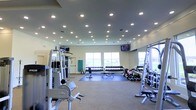 This fitness center is a comprehensive, single-level workout facility designed to accommodate a wide range of exercise needs. The space is illuminated by an abundance of recessed ceiling lights, ensuring a bright and energizing environment throughout. Large windows along one wall invite natural light and provide open views of the surrounding area, contributing to an airy and motivating atmosphere.

The facility is outfitted with a robust selection of professional-grade equipment. Precor strength machines anchor one side of the floor, offering a variety of resistance training options. A full dumbbell rack lines the far wall, featuring an extensive range of free weights neatly organized for easy access, accompanied by exercise benches positioned on protective floor mats. A cable and multi-station weight machine further expands the strength training options available to users. Additional functional training equipment, including medicine balls, rounds out the free weight area.

Multiple flat-screen televisions are mounted at strategic locations throughout the space, allowing exercisers to stay entertained during their workouts. The flooring is a combination of carpeted areas and rubber exercise mats, providing both comfort and safety. The overall layout is open and well-organized, making efficient use of the available floor space while maintaining clear pathways between equipment stations.