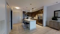 This well-appointed single-floor space showcases a thoughtfully designed open-concept layout that maximizes functionality and flow. The kitchen serves as the centerpiece of the living area, featuring rich dark cabinetry that provides ample storage, paired elegantly with a full suite of stainless steel appliances, including a French door refrigerator, built-in microwave, and a stove. Light-toned countertops offer a striking contrast against the dark cabinetry, creating a balanced and contemporary aesthetic. A generously sized central island anchors the space, complete with a clean white finish and bar-height seating, making it ideal for casual dining or entertaining. Pendant lighting above the island, complemented by a track lighting system, ensures the kitchen is well-lit with a warm, inviting ambiance. The light hardwood flooring extends seamlessly throughout, enhancing the open and airy feel of the space. The entry area is conveniently positioned near the kitchen, featuring a solid door entry. Overall, the space exudes a clean, modern character with a neutral palette, quality finishes, and a practical design that is both stylish and highly functional.