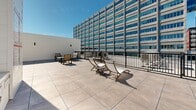 Located atop a residential building at 5480 Avenue Henri-Julien in the Mile-End neighbourhood of Montréal, this rooftop terrace offers a well-appointed outdoor retreat designed for relaxation and socializing. The terrace is finished with large-format porcelain-style tiles laid in a clean grid pattern, giving the space a polished, contemporary aesthetic.

The layout is divided into distinct zones to accommodate a variety of uses. A sun-lounging area features reclining chaise lounge chairs positioned to take full advantage of open sky exposure, ideal for catching rays on clear days. Separate seating clusters, furnished with sturdy slatted chairs and low coffee tables, provide comfortable spaces for conversation and outdoor dining. Wall-mounted light fixtures are integrated into the surrounding parapet walls, ensuring the terrace remains functional into the evening hours.

A central rooftop mechanical enclosure houses stairwell access and utility elements, finished in clean white paneling that maintains a cohesive visual tone throughout the space. The terrace is enclosed by a black metal safety railing along its perimeter, offering unobstructed views of the adjacent Montréal skyline, including a prominent glass-and-concrete office tower. The open sky above and the urban backdrop create a dynamic contrast between the terrace's calm, well-organized design and the vibrant city surrounding it.