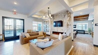 Located in Rosemount, MN, this thoughtfully designed clubroom offers an open-concept layout that seamlessly connects a gourmet kitchen with a sophisticated living area. The kitchen is a standout feature, showcasing bold navy blue cabinetry paired with striking marble-look quartz countertops, a large central island with a dramatic waterfall edge, white subway tile backsplash, wood-look tile flooring, open shelving with warm wood accents, and stylish pendant lighting. Multiple mounted televisions and generous counter space make it ideal for gatherings. The living area exudes elegance with a soaring coffered ceiling, a sculptural multi-globe chandelier, and a full-height marble-clad fireplace wall serving as a dramatic focal point. A wall-mounted television above the linear electric fireplace anchors the space, while plush neutral sofas invite relaxed conversation. A cozy reading nook near large windows provides natural light and a quiet retreat, accented by a curated gallery wall of framed artwork. Deep blue accent walls with decorative shelving add a pop of color and visual interest. Wood-look tile flooring flows consistently throughout, unifying the space with warmth and continuity. This clubroom delivers a refined, contemporary ambiance suited for both social events and casual everyday use.