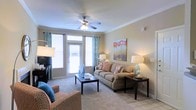This single-floor residence offers a comfortable and thoughtfully designed living environment. The living room serves as the heart of the space, showcasing elegant crown molding along the ceiling line and a classic ceiling fan with integrated lighting. Large windows and a glass-paneled door allow an abundance of natural light to pour in, creating a warm and inviting atmosphere throughout the day. Decorative teal curtains add a pop of color that complements the neutral, warm-toned walls and soft carpeted flooring. The living area provides ample room for relaxed seating arrangements, with space for a sofa, accent chairs, and a central coffee table. Tasteful décor, including table lamps and wall art, enhances the cozy yet polished ambiance. The transition between the carpeted living area and what appears to be tiled flooring near the entryway adds a practical and visually distinct touch. Overall, the space presents a clean, functional layout with a welcoming aesthetic, balancing comfort and style in a bright, naturally lit setting ideal for everyday living.