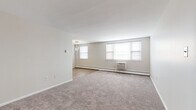 This single-floor residence offers a clean, functional layout designed for comfortable everyday living. The expansive living area is finished with plush wall-to-wall carpeting and neutral-toned walls, creating a bright and welcoming atmosphere. Multiple windows line the far wall, allowing natural light to fill the room, while a wall-mounted air conditioning unit and baseboard heating ensure year-round climate comfort. A small chandelier fixture adds a subtle decorative touch near the entry transition.

The kitchen features an L-shaped layout with light-toned laminate flooring, contrasting white upper cabinetry and dark wood-paneled lower cabinets, and granite-style countertops. It is equipped with a gas cooktop, a wall oven, and a full-size refrigerator, offering practical functionality for home cooking.

The bedroom is generously sized, also carpeted, and includes a built-in closet. A wall-unit air conditioner and baseboard heating mirror the comfort features found in the living area, while a window provides natural light.

The full bathroom showcases a retro-inspired aesthetic with distinctive blue and white tile work on both the walls and mosaic floor. A soaking tub with shower, toilet, and a white vanity with sink complete the space, all tied together by coordinating blue accent fixtures and a wall-mounted mirror.