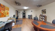 Located within the Business Center at 13401 Morgan Ave S in Burnsville, Minnesota, this single-room office offers a well-organized and versatile workspace designed to support a variety of professional functions. The office features warm hardwood-style flooring throughout, neutral beige walls, and a textured ceiling with flush and overhead lighting fixtures, including ceiling fans that contribute to a comfortable working environment.

The space is thoughtfully arranged into distinct functional zones. A workstation area accommodates multiple glass-top desks paired with ergonomic task chairs, each equipped with computer monitors — ideal for day-to-day administrative or collaborative work. Area rugs beneath the desks define the workspace and add a layer of comfort underfoot.

A round conference table with a star-motif surface and accompanying chairs provides a dedicated meeting or consultation area, lending the office a personable and professional character. Bookshelves and corner display units line the walls, stocked with books and decorative items, adding both utility and warmth to the space.

Artwork adorns the walls, including a vibrant large-scale canvas painting and decorative metal wall pieces, giving the office an inviting and creative atmosphere. A solid entry door with a narrow sidelight panel provides access and a measure of privacy. The overall layout makes efficient use of the available space, supporting both focused individual work and small group interaction.