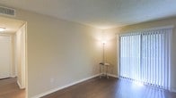 This single-floor space presents a clean, straightforward layout designed for comfortable everyday living. The main living area is finished with dark hardwood-style flooring that contrasts attractively against the warm, neutral-toned walls, creating a grounded and cohesive aesthetic. A textured popcorn ceiling adds a classic architectural detail overhead. A sliding glass door fitted with vertical blinds serves as a prominent feature, allowing natural light to filter into the room while offering easy access to the outdoors. The overall color palette — soft beiges and warm whites — lends the space a calm, understated ambiance. White baseboards provide a crisp finish along the perimeter of the room, complementing the light window framing. The open wall space offers flexibility for a variety of furniture arrangements, making the area adaptable to different uses. The combination of natural light from the sliding door and the simplicity of the finishes gives the space a bright, welcoming character suited for a range of lifestyles.
