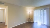 This single-floor space presents a clean, straightforward layout designed for comfortable everyday living. The main living area is finished with dark hardwood-style flooring that contrasts attractively against the warm, neutral-toned walls, creating a grounded and cohesive aesthetic. A textured popcorn ceiling adds a classic architectural detail overhead. A sliding glass door fitted with vertical blinds serves as a prominent feature, allowing natural light to filter into the room while offering easy access to the outdoors. The overall color palette — soft beiges and warm whites — lends the space a calm, understated ambiance. White baseboards provide a crisp finish along the perimeter of the room, complementing the light window framing. The open wall space offers flexibility for a variety of furniture arrangements, making the area adaptable to different uses. The combination of natural light from the sliding door and the simplicity of the finishes gives the space a bright, welcoming character suited for a range of lifestyles.