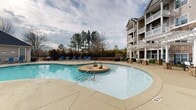Located at 6443 Zebulon Rd in Macon, Georgia, this two-floor residential community is designed with a strong emphasis on outdoor amenity spaces that complement its traditional architectural style. The multi-building complex features neutral-toned siding, stone accents, pitched rooflines, and private balconies that overlook the well-appointed grounds below.

At the heart of the outdoor area is a generously sized, freeform swimming pool with clear blue water, entry steps, and a central island water feature. The expansive concrete pool deck is furnished with blue lounge chairs, providing ample space for sunbathing and relaxation. A dedicated pool house with large windows serves as an additional amenity structure adjacent to the main pool, offering interior space with direct views of the pool area.

A white pergola structure positioned along the pool perimeter provides shaded seating areas with outdoor dining tables and stone pillar accents, creating an ideal setting for outdoor gatherings. A standout feature of the grounds is a substantial stone outdoor fireplace, adding a refined, communal focal point to the landscaped exterior. Iron fencing encloses the pool and amenity areas, while mature trees and manicured shrubbery contribute to the overall setting.