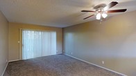 This single-floor space presents a straightforward and functional layout centered around a generously sized living area. The room features wall-to-wall carpet in a neutral tone, providing a comfortable and cohesive foundation throughout the space. Warm, beige-toned walls create an inviting atmosphere, complemented by clean white baseboards that frame the room neatly.

A ceiling fan with an integrated light fixture is centrally positioned, offering both ambient lighting and air circulation. The textured popcorn ceiling adds a classic residential character to the space. A large sliding glass door, dressed with full-length vertical blinds, serves as the focal point of one wall, allowing a generous amount of natural light to filter into the room while offering privacy control. The blinds diffuse the incoming light softly, contributing to the room's overall warmth.

Electrical outlets are conveniently placed along the walls, allowing for flexible furniture arrangement. The overall ambiance is calm and neutral, making the space a practical canvas well-suited for a variety of interior styles and furnishing preferences.