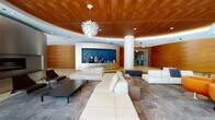 Located at 127 N Dearborn Street in the heart of Chicago's Loop, this single-floor amenity lounge delivers a refined and contemporary atmosphere designed for comfort and collaboration. The space is anchored by a striking curved wood-paneled ceiling with integrated cove lighting, which creates a warm, organic contrast against the clean white structural columns and light-toned tile flooring throughout.

The lounge is organized into several distinct seating zones, offering both intimate and open gathering options. Plush white leather sectional sofas, modular upholstered benches, and vibrant orange swivel chairs provide a variety of seating arrangements suited to casual meetings or relaxed socializing. A sleek linear fireplace set into a brushed metal wall panel adds a focal point to one side of the space, enhancing its inviting ambiance.

A dedicated beverage station features a blue mosaic tile backsplash and professional coffee equipment, serving as a functional and visually dynamic amenity. Floor-to-ceiling glass walls on one side of the lounge flood the interior with natural light and offer views of outdoor greenery, seamlessly connecting the interior to the exterior environment. The entry corridor is finished with rich wood-toned wall panels and polished tile, setting an elevated tone from arrival. A sculptural pendant light fixture serves as a central design statement, completing this thoughtfully appointed space.