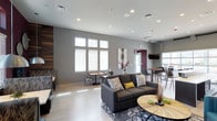 This single-floor amenity space is designed with a contemporary aesthetic, offering a versatile and inviting environment for socializing and relaxation. The open-concept layout is anchored by a spacious lounge area featuring a plush charcoal sectional sofa, a patterned area rug, and a sleek round wood coffee table. Vibrant teal leather accent chairs provide a bold pop of color against the cool gray walls, creating a dynamic and modern atmosphere.

A dedicated seating nook, set against a rich burgundy feature wall, offers cozy booth-style banquette seating beneath elegant pendant lights and decorative wall medallions. A wall-mounted flat-screen TV serves this area, adding an entertainment focal point. Additional high-top table seating and bar-style chairs are available for more casual gatherings.

A striking vertical wood-plank accent wall adds warmth and texture, contrasting beautifully with the light hardwood-style flooring that runs throughout the space. Natural light pours in through multiple large windows fitted with white blinds, enhancing the bright and airy feel.

The kitchenette area is outfitted with dark cabinetry, light quartz countertops, stainless steel appliances, and a utility sink. A large roll-up garage-style glass door opens the kitchenette directly to an outdoor area, seamlessly blending indoor and outdoor functionality. Recessed lighting throughout ensures a well-lit, welcoming environment.