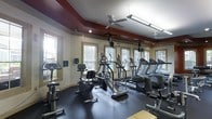 This fitness center is designed to support a comprehensive cardio-focused workout experience. The room is lined with multiple large windows along two walls, allowing generous amounts of natural light to fill the space and offering views of the surrounding outdoor greenery. The flooring is a durable dark rubber material, ideal for high-traffic exercise environments.

The equipment layout is thoughtfully organized, featuring a row of treadmills positioned along the windowed wall, complemented by elliptical trainers, stair climbers, stationary bikes, and a recumbent cycle. Mounted televisions are strategically placed throughout the room, allowing users to stay entertained during workouts.

The ceiling is a standout architectural detail, featuring a two-tone design with a deep burgundy tray accent that frames the upper perimeter of the room. Ceiling fans are installed to maintain airflow and comfort during exercise. Recessed and track lighting provides additional illumination during low-light conditions.

Wall-mounted fitness accessories and informational panels are visible near the entrance, adding functional value. Overall, the space presents a clean, organized, and energizing environment suited for daily fitness use.